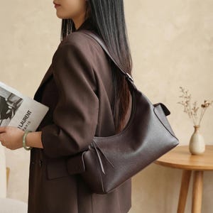 Puede incluir: Un bolso de hombro de cuero marrón oscuro con un diseño curvo. El bolso tiene una sola correa y un pequeño bolsillo lateral. El bolso se lleva sobre el hombro. La imagen también muestra una revista con el texto "LAURENT PARIS".