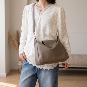 May include: A taupe leather shoulder bag with a flap closure and a long, adjustable strap. The bag has a simple, minimalist design and is worn over the shoulder. The bag is made of a soft, supple leather material.