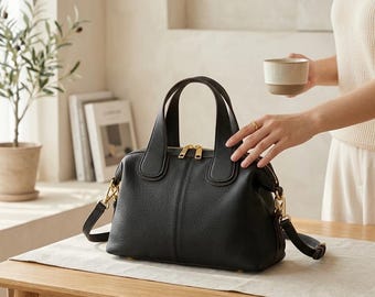 May include: A black leather handbag with gold-coloured zips and a detachable shoulder strap. The bag has a structured design with a top handle. It is placed on a wooden table. A small plant and books are in the background.