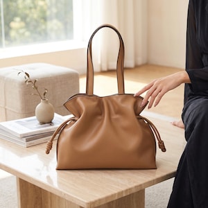 May include: A tan shoulder bag featuring a soft, gathered silhouette with a drawstring closure on each side. The bag includes two long, thin handles and is constructed from a smooth, solid-colored material with knotted ends on the drawstrings.