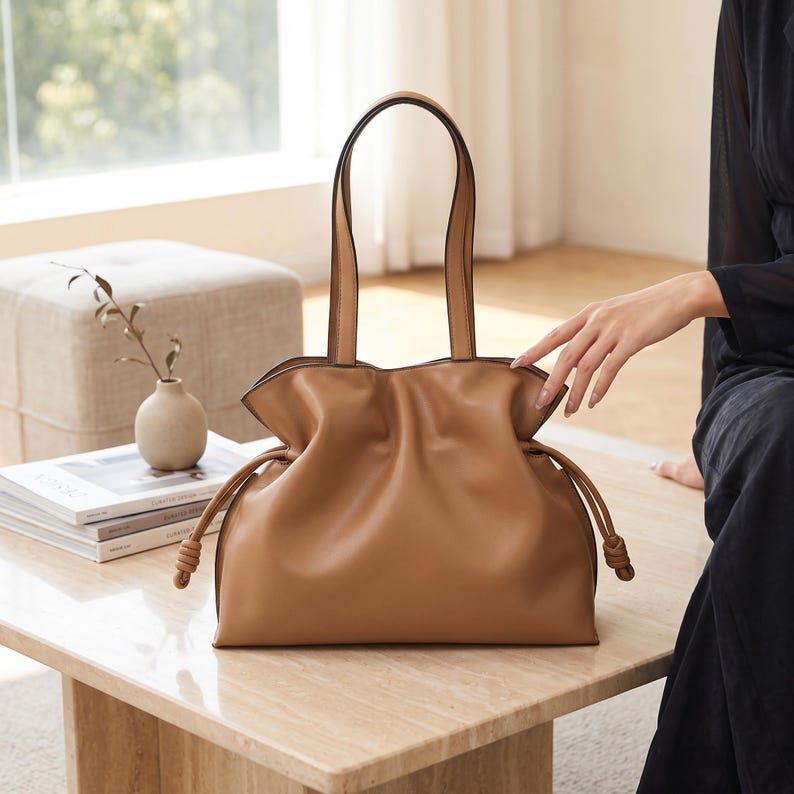 May include: A tan shoulder bag featuring a soft, gathered silhouette with a drawstring closure on each side. The bag includes two long, thin handles and is constructed from a smooth, solid-colored material with knotted ends on the drawstrings.