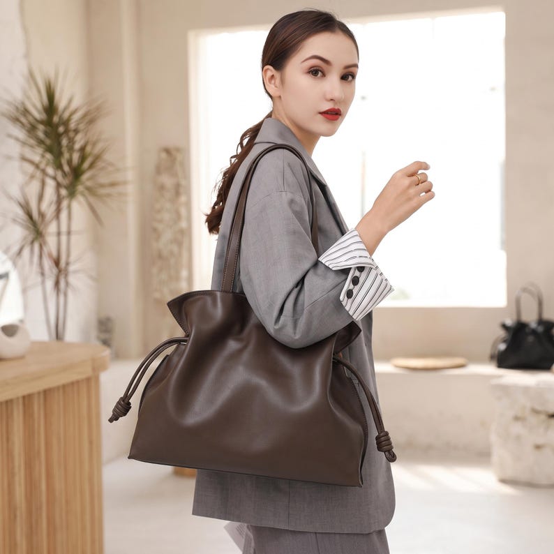 May include: A dark brown shoulder bag with a smooth finish, featuring a soft, slouchy silhouette, a wide main compartment, and thin side drawstrings that end in knotted details. The bag has two long, slender shoulder straps for carrying.