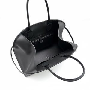 May include: A black leather tote bag with two long handles. The bag has a spacious interior with a zipped pocket and a smaller pocket. The bag is open, revealing the interior lining and construction. The bag is designed for carrying items.