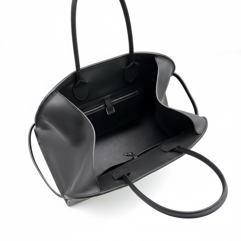 May include: A black leather tote bag with two long handles. The bag has a spacious interior with a zipped pocket and a smaller pocket. The bag is open, revealing the interior lining and construction. The bag is designed for carrying items.