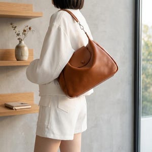 May include: A tan leather shoulder bag with a single strap and a button closure. The bag is a warm, medium tan colour and appears to be made of soft leather. The bag is being carried by a person wearing a white jacket and shorts.