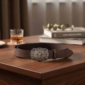 May include: A brown leather belt with a decorative silver buckle featuring a bull's head. The belt has a textured pattern. The buckle is the focal point, adding a rustic touch. The belt is displayed on a wooden surface.