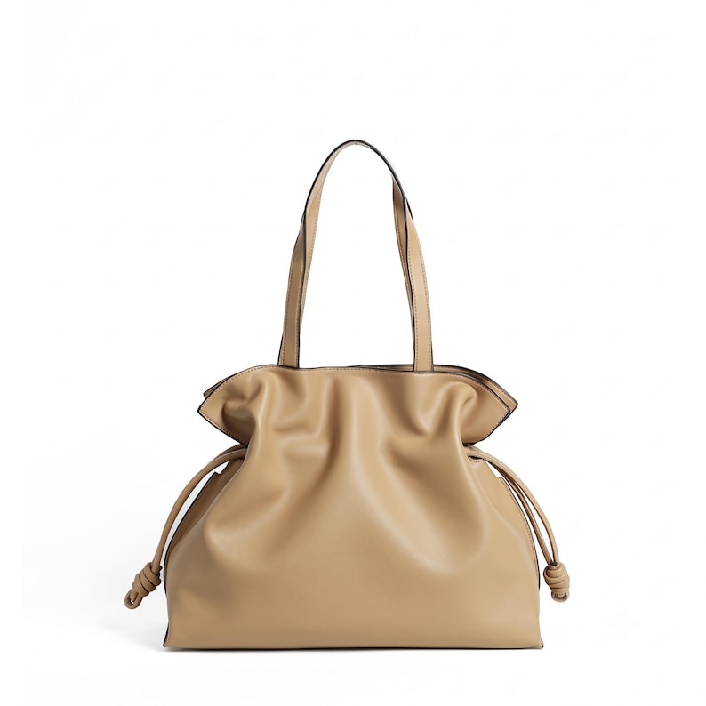 May include: A beige tote bag featuring a soft, gathered silhouette with a drawstring closure on both sides. The bag includes two long shoulder straps and knotted ends on the drawstrings, creating a relaxed, slouchy shape with smooth texture.