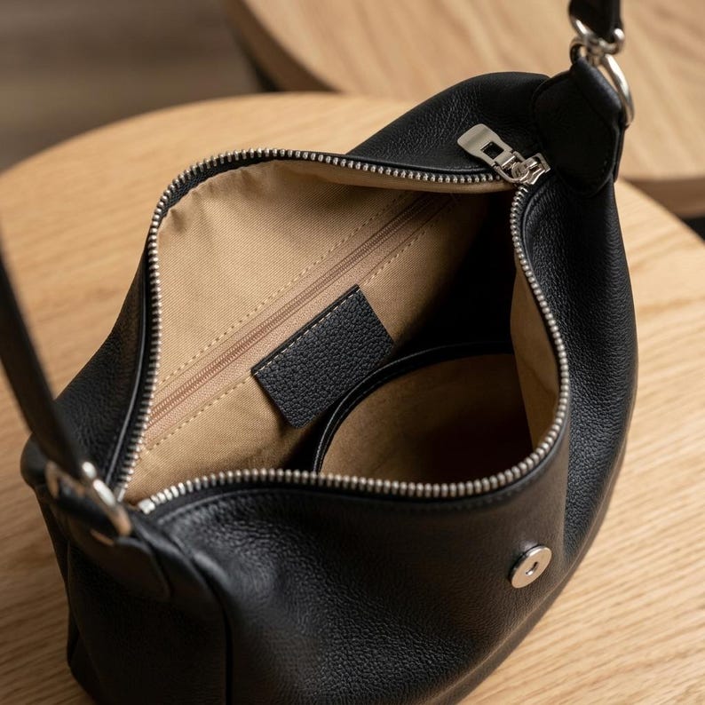 May include: Black leather shoulder bag with a silver zip and fittings. The bag is open, showing a tan interior and a small pocket. The bag has a curved shape and a strap for carrying over the shoulder.