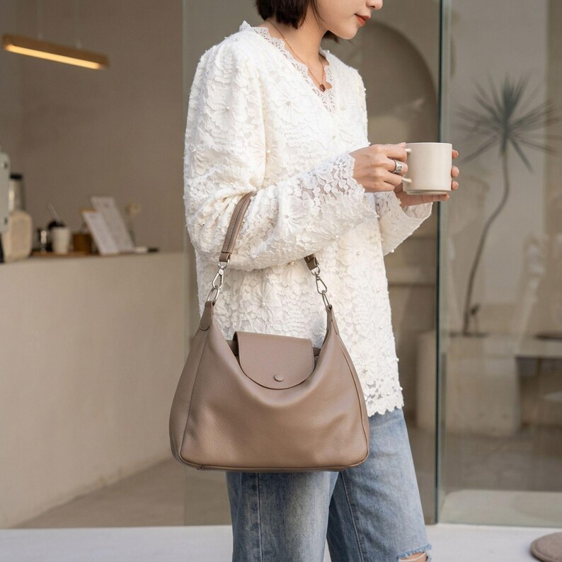 May include: A taupe leather shoulder bag with a flap closure and a silver chain strap. The bag is crescent-shaped and has a large capacity. The woman is wearing a white lace top and blue jeans.