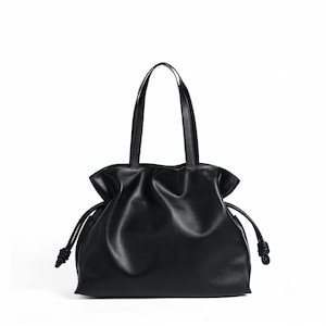 May include: A black tote bag with a smooth finish, featuring a gathered top opening and two thin shoulder straps. The sides of the bag include drawstring cords that end in knotted details, creating a soft, pleated silhouette throughout the body.