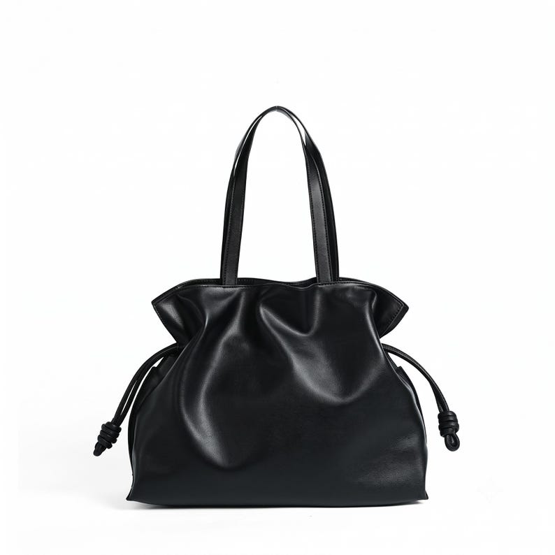 May include: A black tote bag with a smooth finish, featuring a gathered top opening and two thin shoulder straps. The sides of the bag include drawstring cords that end in knotted details, creating a soft, pleated silhouette throughout the body.