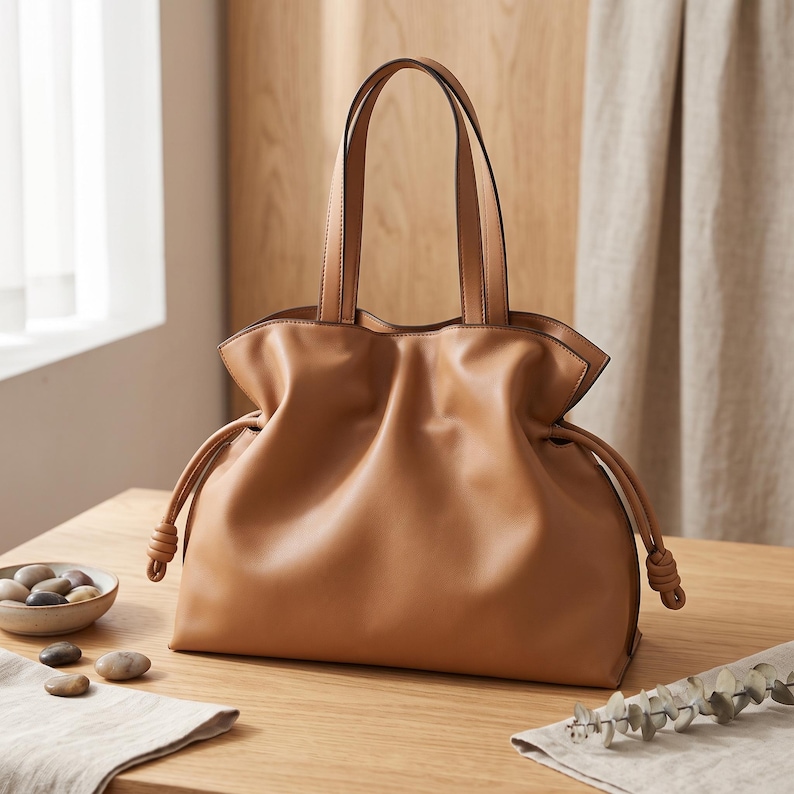May include: A tan leather tote bag featuring a soft, slouchy silhouette with a gathered top opening. The bag includes two vertical shoulder straps and side drawstrings that terminate in knotted ends, creating a pleated effect along the sides.