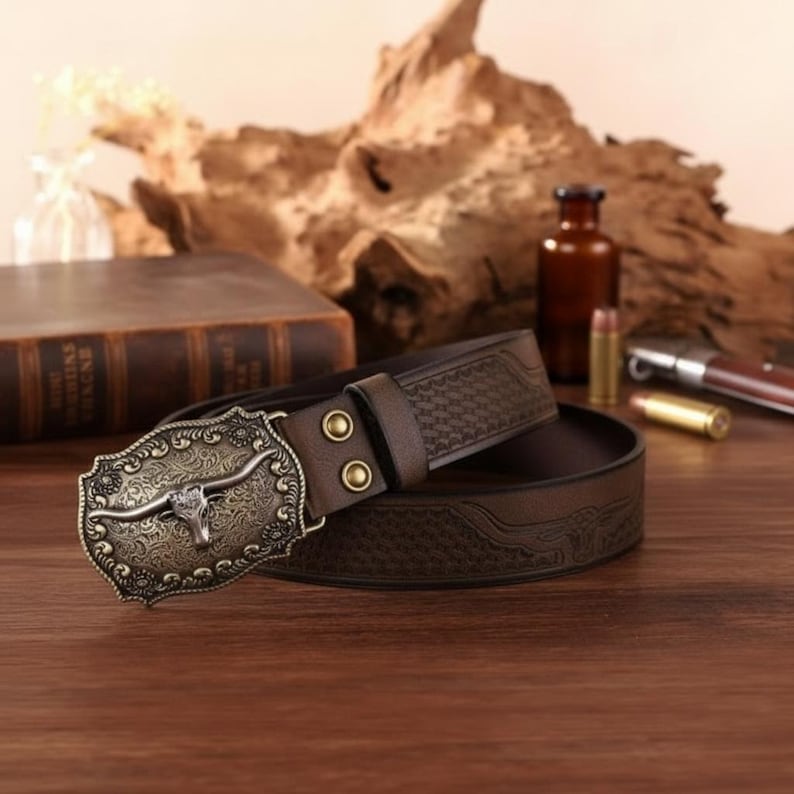 May include: Brown leather belt with a decorative metal buckle featuring a bull skull design. The belt has a textured pattern and two brass-coloured rivets. The buckle is antique-finished.