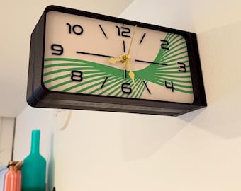 May include: A rectangular wall clock with a black frame and a white face featuring black numerals 2, 3, 4, 6, 8, 9, 10, and 12. The clock face displays a green radiating line pattern and has gold-colored hour, minute, and second hands.