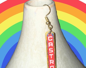 May include: A single gold-colored earring with the word "CASTRO" in red letters. The earring hangs from a gold hook. The background features a rainbow arc and a white vase.