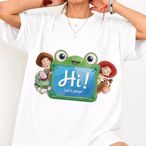 May include: A white short-sleeve t-shirt featuring a graphic of Toy Story characters surrounding a green frog-shaped tablet. The tablet screen displays the text Hi! Let's play! The characters include Woody, Buzz Lightyear, Jessie, and Bullseye.