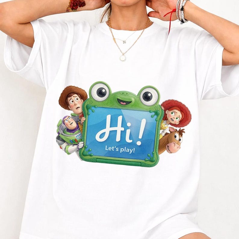 May include: A white short-sleeve t-shirt featuring a graphic of Toy Story characters surrounding a green frog-shaped tablet. The tablet screen displays the text Hi! Let's play! The characters include Woody, Buzz Lightyear, Jessie, and Bullseye.