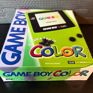 May include: A boxed Game Boy Color video game console in kiwi green. The box features the words "GAME BOY COLOR" in various colors and fonts. The console's screen is visible on the box, along with the Nintendo logo.