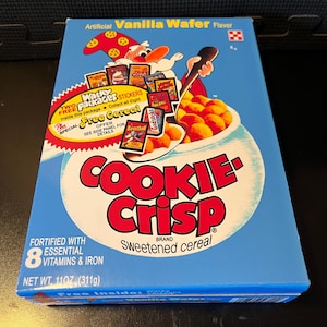 May include: A blue Cookie Crisp cereal box with a cartoon chef holding a spoon of cereal. The box features the text "Cookie Crisp" and "Sweetened cereal". It contains 11 oz (311g) of cereal and includes free stickers.