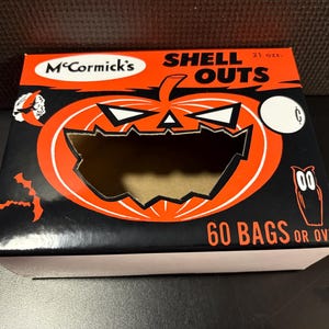 May include: A rectangular McCormick's Shell Outs box with a black and orange Halloween theme. The front features a large jack-o'-lantern cutout, a witch on a crescent moon, an owl, and bats. Text reads: McCormick's, Shell Outs, 21 ozs., 60 bags or over.