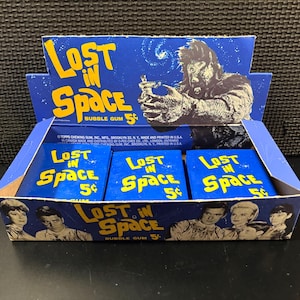 May include: A vintage display box of "Lost in Space" bubble gum. The box is blue and yellow, featuring images of characters and a monster. Each pack of gum is 5 cents.