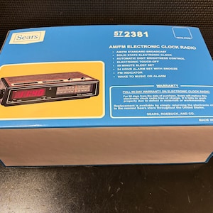May include: A vintage Sears AM/FM electronic clock radio in its original box. The box is blue with a yellow rectangle displaying the clock radio. The clock radio has a wood-grain top, a digital display, and a black frame. Text on the box includes "AM/FM Electronic Clock Radio."