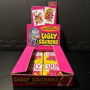 May include: A pink cardboard display box for "Ugly Stickers" bubble gum. The box features the product name in large yellow letters, with monster graphics. The display includes individual packs of gum and promotional cards.