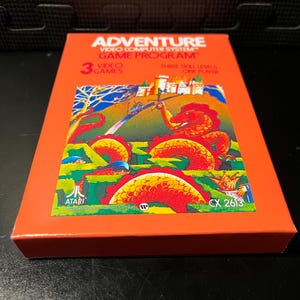 May include: An orange Atari Adventure video game program box. The box features the game title and a colorful illustration of a dragon, castle, and landscape. The box reads "3 Video Games" and "CX 2613".