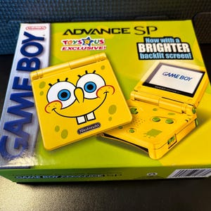 May include: A yellow Game Boy Advance SP console featuring a SpongeBob SquarePants design. The box reads "TOYS R US EXCLUSIVE!" and "Now with a BRIGHTER backlit screen!" The console is yellow with green buttons and a SpongeBob face.