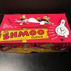 May include: A red box featuring the text "Lux Shmoo Clock" in yellow. The box has cartoon illustrations of people and a clock shaped like a white creature. The text "The up-to-the-minute Shmoo right on time for you!" is also visible.