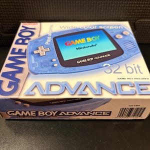 May include: A light blue Game Boy Advance box with the words "GAME BOY ADVANCE" in large, bold letters. The box features an image of the handheld console with a wide color screen. The text "32 bit" is also visible.
