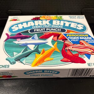 May include: A box of Shark Bites chewy fruit snacks. The box features colorful shark illustrations in various colors, including red, blue, and yellow. The text on the box reads "Shark Bites" and "Fruit Punch". The net weight is 5.4 oz.