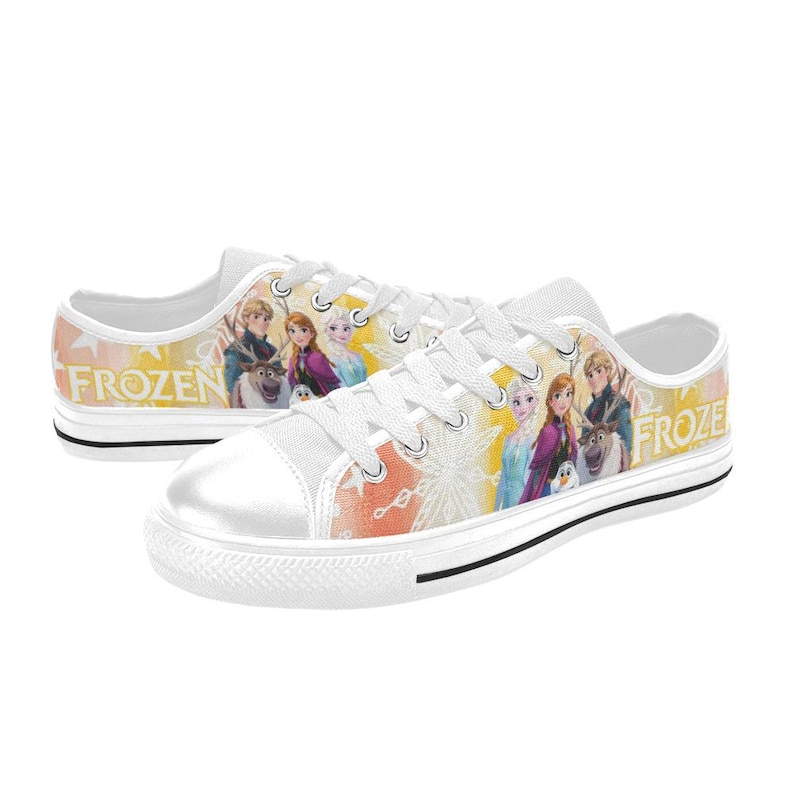 Puede incluir: Zapatillas de lona blancas con personajes de la pel&iacute;cula de animaci&oacute;n "Frozen". Los zapatos tienen una puntera blanca, cordones blancos y una franja negra a lo largo de la suela. La palabra "FROZEN" est&aacute; impresa en amarillo en el lateral.