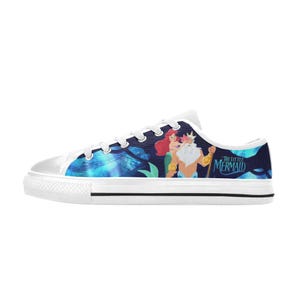 May include: White canvas sneakers featuring a colorful design from "The Little Mermaid." The design includes Ariel, King Triton, and the movie's title. The shoes have a white rubber sole and laces.