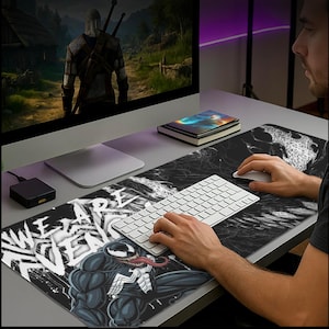 Może przedstawiać: Czarna i biała przedłużona podkładka pod mysz z postacią Venom z Marvel Comics. Podkładka ma biały napis "WE ARE VENOM", z czarno-białym marmurowym wzorem po prawej stronie. Klawiatura i mysz są na niej.