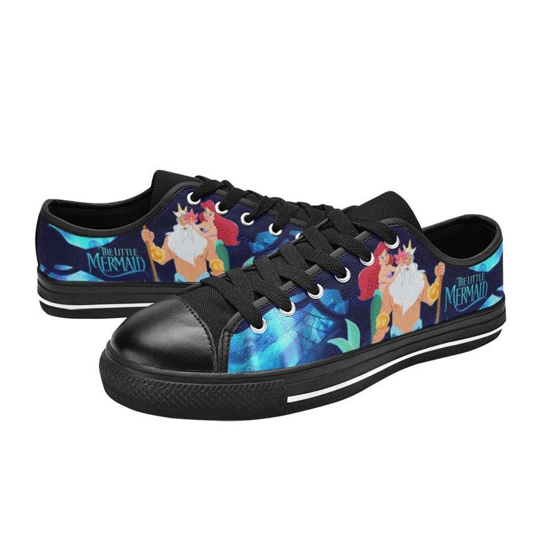 May include: Black canvas sneakers featuring a colorful illustration of characters from "The Little Mermaid." The design includes King Triton, Ariel, and the movie's logo. The shoes have black laces, a black toe cap, and a white midsole.