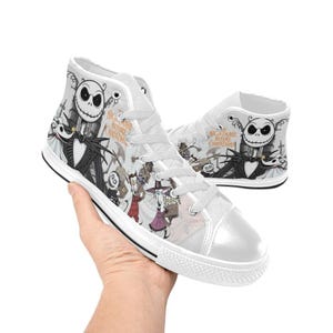 Puede incluir: Zapatillas altas blancas con un estampado colorido de personajes de "Pesadilla antes de Navidad". El dise&ntilde;o incluye a Jack Skellington, Sally y otras figuras. Tambi&eacute;n es visible el texto "Nightmare Before Christmas".