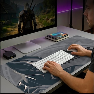 May include: A large, rectangular desk mat with a dark gray and blue design featuring a stylized image. A white keyboard and mouse are on the mat. A computer monitor displays a video game scene.