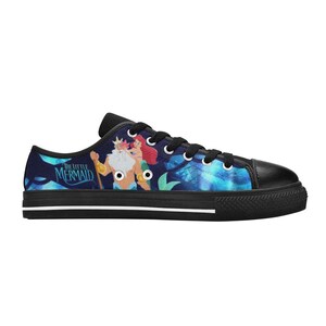 May include: Low-top canvas sneakers featuring a colorful illustration from "The Little Mermaid." The design includes King Triton, Ariel, and a blue and green underwater scene. The shoes have black laces, a black toe cap, and a black sole.