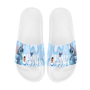 Puede incluir: Sandalias deslizantes blancas con una banda azul que presenta a Stitch de Lilo & Stitch. La banda muestra la palabra "Ohana" e im&aacute;genes de dibujos animados de Stitch. Estas sandalias est&aacute;n dise&ntilde;adas para uso casual.