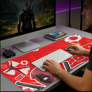 May include: A red and white Deadpool-themed desk mat with a keyboard and mouse. The mat features Deadpool's face, logo, and other graphics. A computer monitor displays a video game scene.