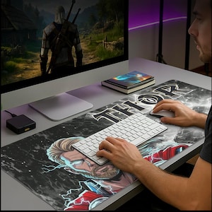 May include: A black and gray rectangular mousepad featuring a graphic of Thor with lightning bolts and the word "THOR" in white. A white keyboard and mouse are on the pad. A computer monitor displays a video game scene.
