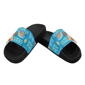 Puede incluir: Un par de sandalias deslizantes negras con una correa azul con la palabra "Moana" y varios dise&ntilde;os tropicales. Las sandalias tienen una suela blanca y est&aacute;n dise&ntilde;adas para uso casual.