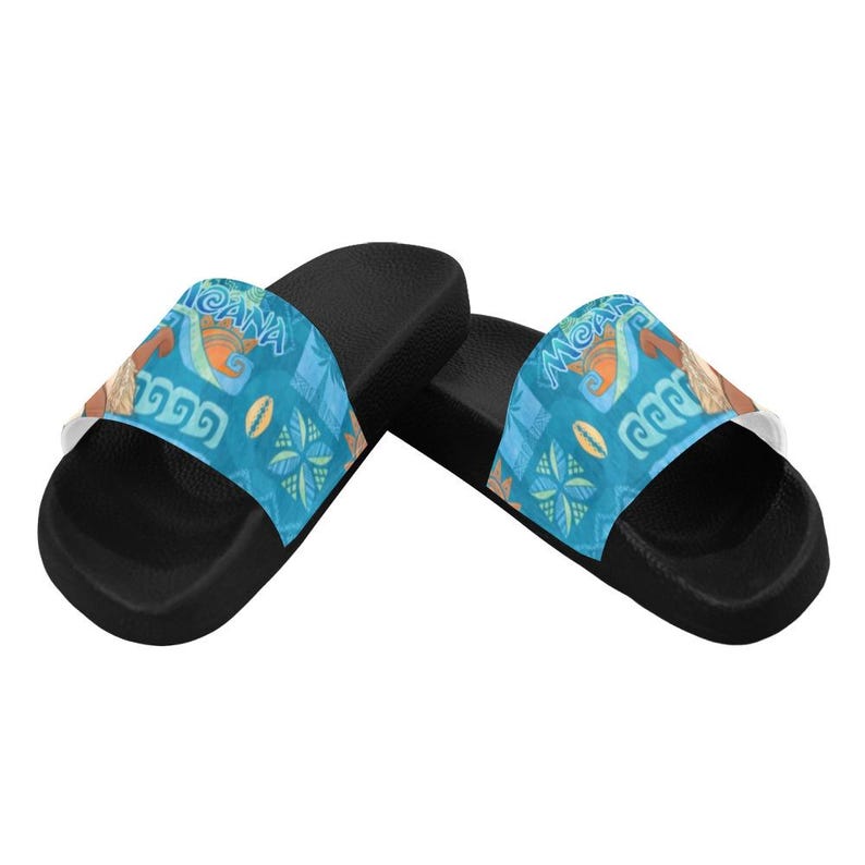 Puede incluir: Un par de sandalias deslizantes negras con una correa azul con la palabra "Moana" y varios dise&ntilde;os tropicales. Las sandalias tienen una suela blanca y est&aacute;n dise&ntilde;adas para uso casual.