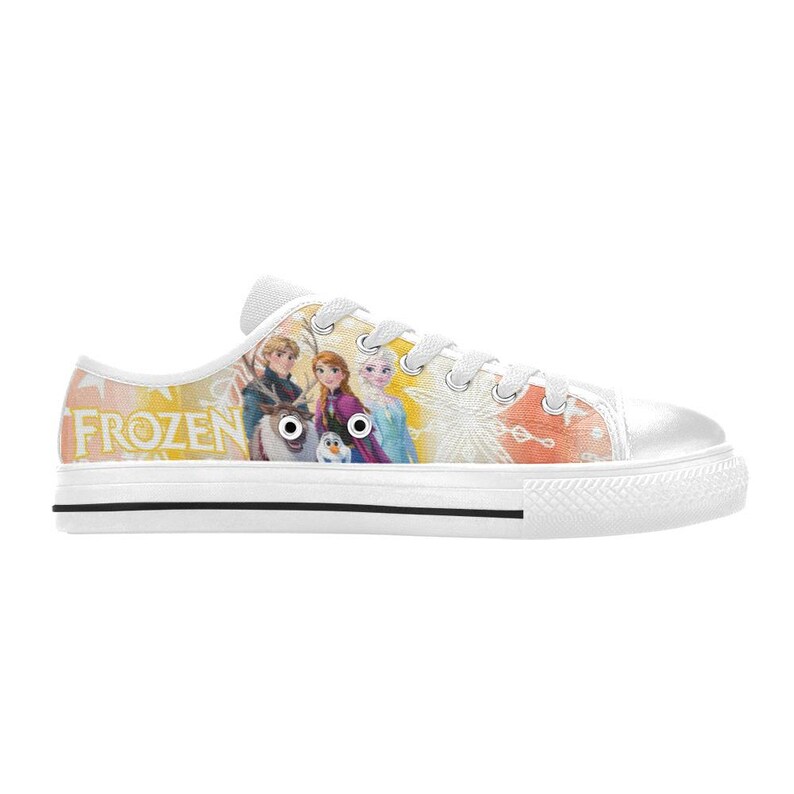 Puede incluir: Zapatillas de lona blancas con personajes de la pel&iacute;cula "Frozen." El dise&ntilde;o incluye a Elsa, Anna, Kristoff y Olaf, con la palabra "FROZEN" en amarillo. Los zapatos tienen cordones blancos y una franja negra en la suela.