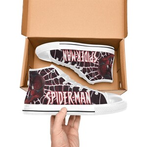 May include: High-top canvas sneakers featuring a Spider-Man design. The shoes are white with a black and red web pattern and the words "SPIDER-MAN" in white. The shoes are in a cardboard box.