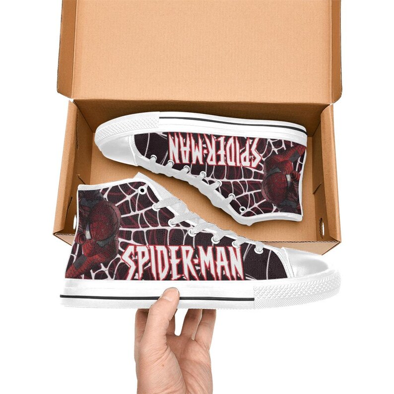 May include: High-top canvas sneakers featuring a Spider-Man design. The shoes are white with a black and red web pattern and the words "SPIDER-MAN" in white. The shoes are in a cardboard box.