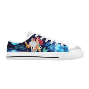 May include: White canvas sneakers featuring a colorful illustration of characters from "The Little Mermaid." The design includes King Triton, Ariel, and the movie title. The shoes have white laces and a black stripe on the sole.