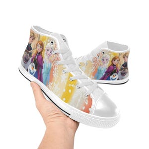 May include: White high-top sneakers featuring a colorful design with characters from the animated movie Frozen. The shoes have a white rubber sole, white laces, and a vibrant print of Elsa, Anna, Olaf, and other characters.