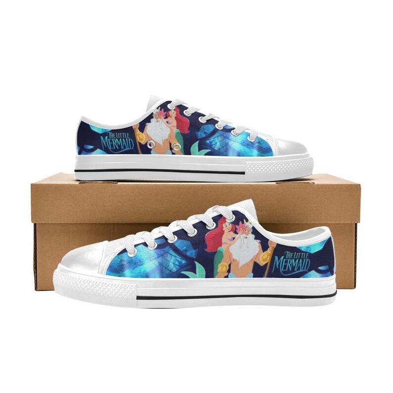 May include: White-soled canvas sneakers featuring a design from "The Little Mermaid." The shoes have a blue and teal background with illustrations of characters and the movie title. The sneakers are displayed on a cardboard box.